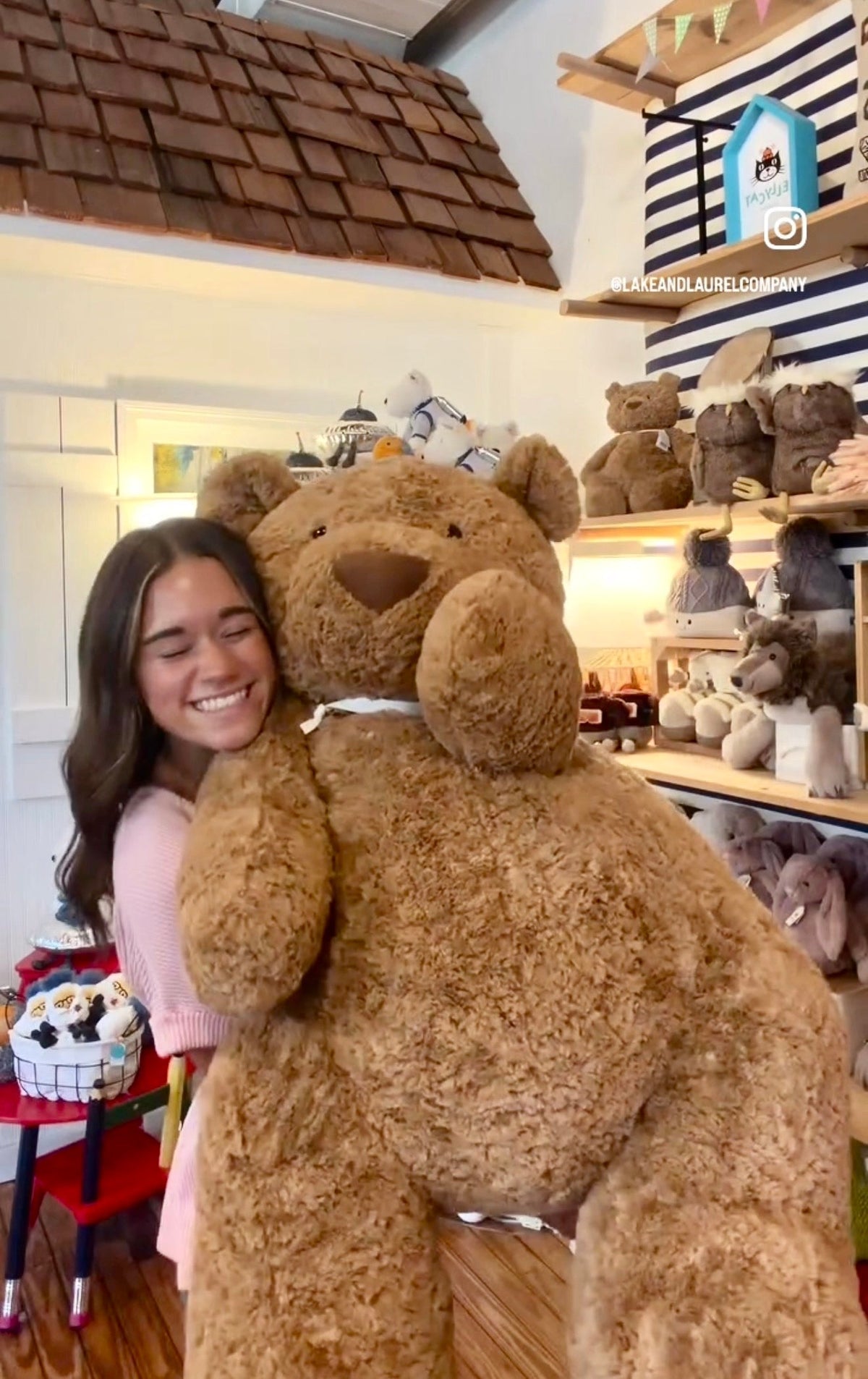 A smiling person hugging a gigantic brown Jellycat Bartholomew Bear in the Lake and Laurel shop
